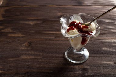 Beyaz vanilyalı dondurma topları ile ahududu reçeli tatlı bardağında kaşık. Kaba ahşaptan yapılmış eski bir masada süslenmiş dondurma. Kopyala-uzayı düzlemi.