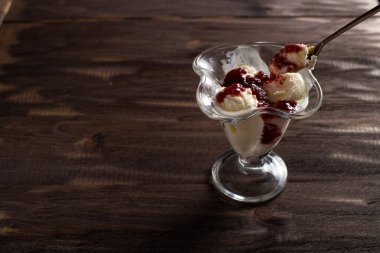Beyaz vanilyalı dondurma topları ile ahududu reçeli tatlı bardağında kaşık. Kaba ahşaptan yapılmış eski bir masada süslenmiş dondurma. Kopyala-uzayı düzlemi.
