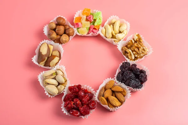 A serving of assorted candied fruit, dried cherries, almonds, raisins, walnuts and hazelnuts in paper muffin cups on a pink background. In the shape of a ring.