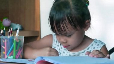 Evdeki pencerenin yanındaki masada oturan, kağıt üzerine resim yapmak için renkli kalemler kullanan sevimli Asyalı küçük kız. Anaokulu öğrencileri Corona virüsü salgını sırasında evde çalışıyorlar..