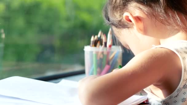 紙に色鉛筆で絵を描くかわいいアジアの女の子は 家の窓のそばのテーブルに座っています コロナウイルス流行時に自宅で勉強する未就学児 — ストック動画