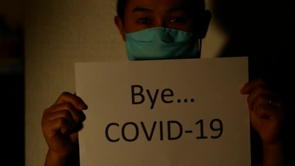 ライトアジアの女性に別れCovid 19というラベルの紙を保持輝いて 白い背景に立つ コロナウイルスのパンデミックにお別れを言う概念 — ストック動画