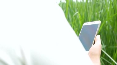 Genç kadın çiftçinin elinde akıllı telefonuyla mısır tarlasının yanındaki çim biçme bıçağını incelerken..