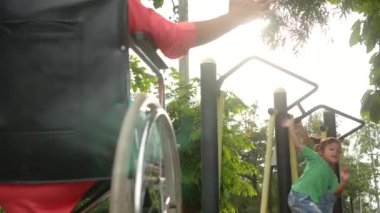 Mutlu engelli kadın tekerlekli sandalyede oturuyor ve kızını açık hava parkında egzersiz malzemeleriyle oynarken izliyor..