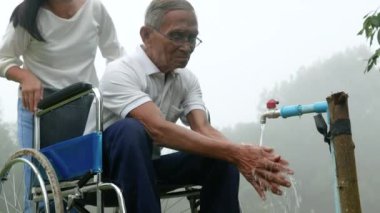 Asyalı bakıcı torun büyükbabasına bakıyor. Parkta seyahat ederken tekerlekli sandalyede oturuyor..