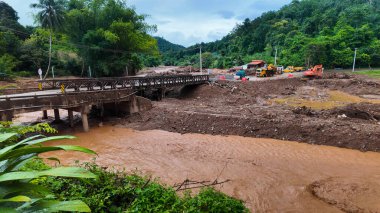 Şiddetli selden sonra çamurlu bir nehri geçen köprü toprak erozyonu, enkaz ve kırsal bölgedeki hasarlı çevreyi gösteriyor..