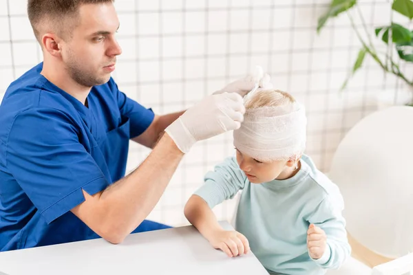 Doctor in blauw uniform aanraken van een jongen hoofd met trauma in zijn hoofd en elastische verbonden rond zijn hoofd. — Stockfoto