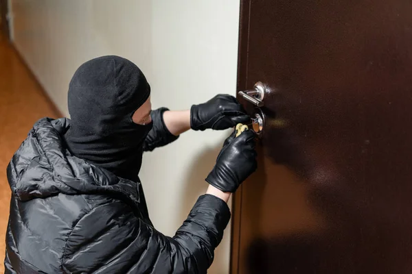 Thief in black mask trying to pick lock a door. House thief concept.