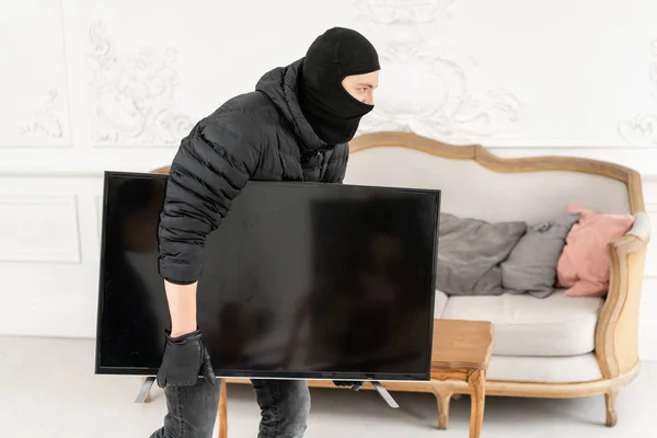 Voleur avec cagoule noire qui vole la télévision moderne et chère. Appartement de luxe avec stuc. Homme cambrioleur voler le téléviseur de la maison — Photo