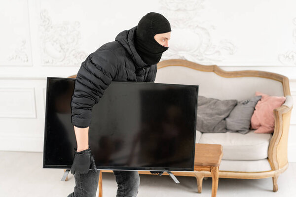 Thief with black balaclava stealing modern expensive television. Luxury apartment with stucco. Man burglar stealing tv set from house