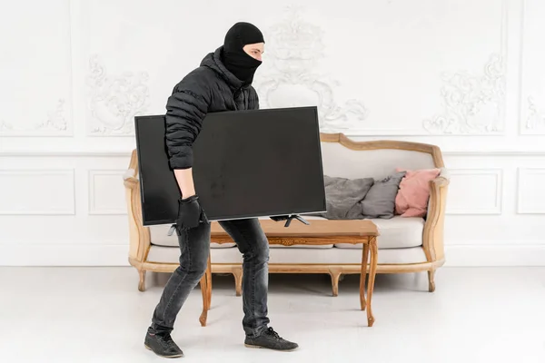 Thief with black balaclava stealing modern expensive television. Luxury apartment with stucco. Man burglar stealing tv set from house