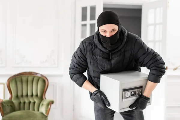 Tjuv tittar på kameran med svart Balaclava stjäla modern elektronisk Safe box. Inbrottstjuv begår ett brott i Lyxlägenhet med stuckatur. — Stockfoto