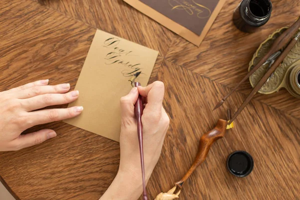 O Calígrafo está a segurar uma caneta com tinta. A menina escreve uma carta em fonte manuscrita. Oficina criativa do artista. Foco suave . — Fotografia de Stock