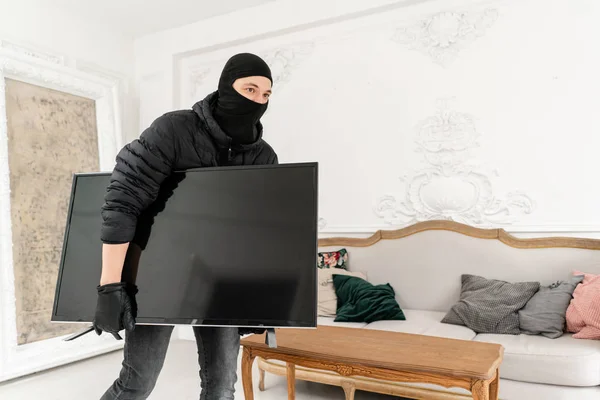 Un ladro che ruba un televisore da casa. Ladro con passamontagna nero rubare moderna televisione costosa. Appartamento di lusso con stucco . — Foto Stock