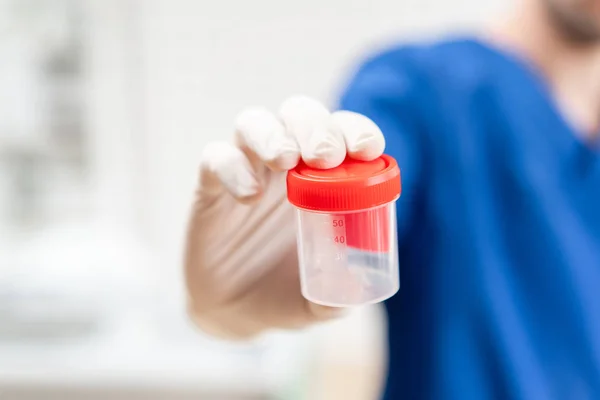 Médecin en uniforme bleu et gants en latex tient un récipient en plastique vide pour prélever des échantillons d'urine, fond clair. Concept médical . — Photo