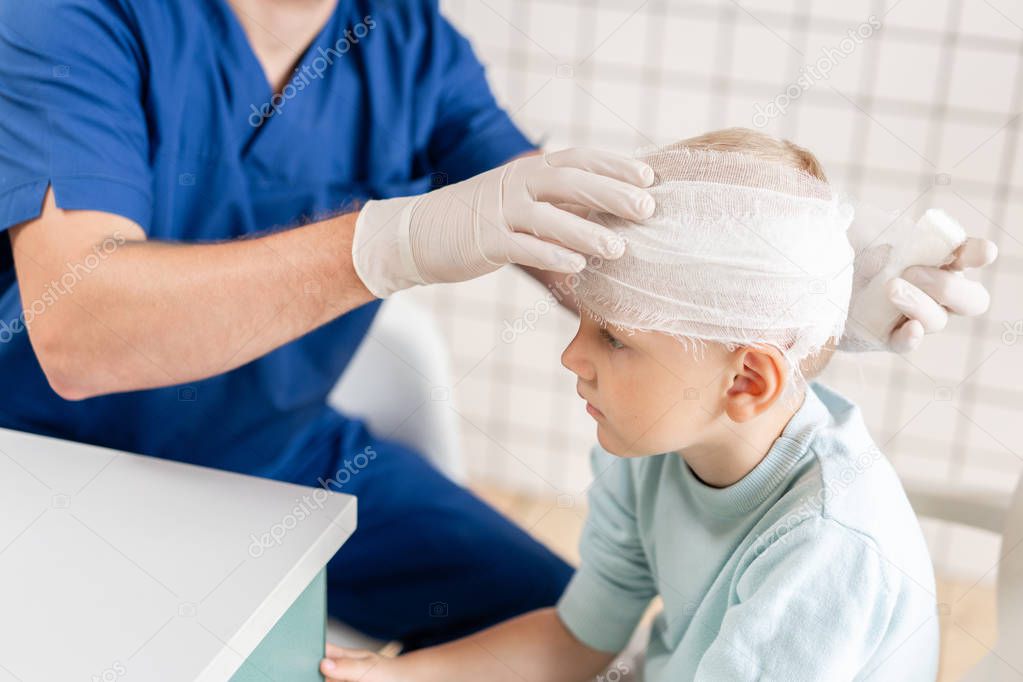 Doctor in blue uniform touching a boy head with trauma in his head and elastic bandaged around his head.