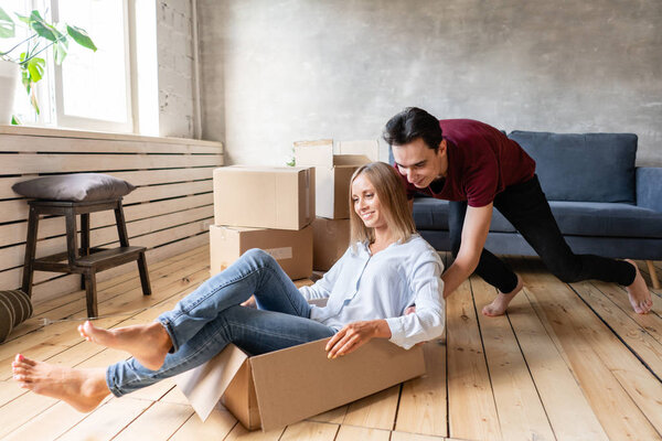 Happy couple having fun and riding in cardboard boxes at new home. Young couple moving to a new apartment together. Relocation concept