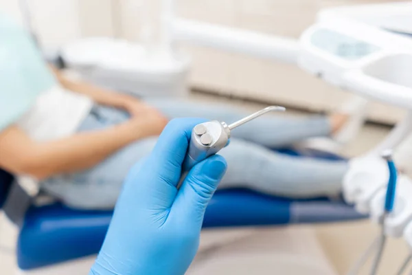 Κοντινό-up χέρι του οδοντίατρου στο γάντι κρατά όπλο οδοντιατρικό νερό και αέρα. Ο ασθενής σε μπλε καρέκλα στο παρασκήνιο. Γραφείο όπου ο οδοντίατρος διεξάγει επιθεώρηση και. — Φωτογραφία Αρχείου