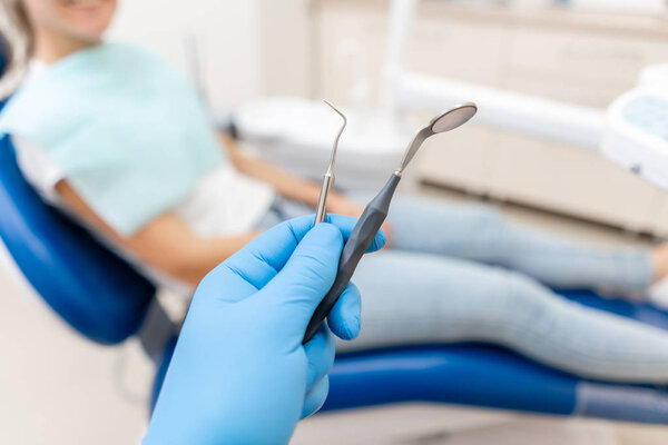 Close-up hand of dentist in the glove holds tool probe and mirror. The patient in the dental chair at the background. Dental work in clinic. Office where dentist conducts inspection and concludes.