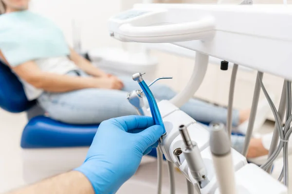 Κοντινό χέρι του οδοντίατρου στο γάντι κρατά οδοντοστρόβιλο υψηλής ταχύτητας. Ο ασθενής σε μπλε καρέκλα στο παρασκήνιο. Γραφείο όπου ο οδοντίατρος διεξάγει επιθεώρηση και. — Φωτογραφία Αρχείου