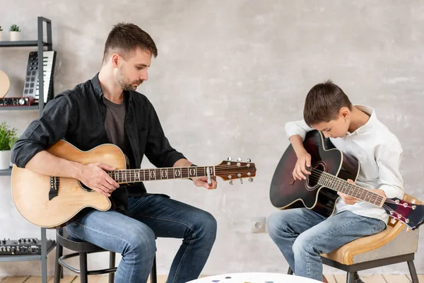 Two brothers learn to play acoustic guitar. The older brother tunes the guitar while the younger one runs his finger along the strings