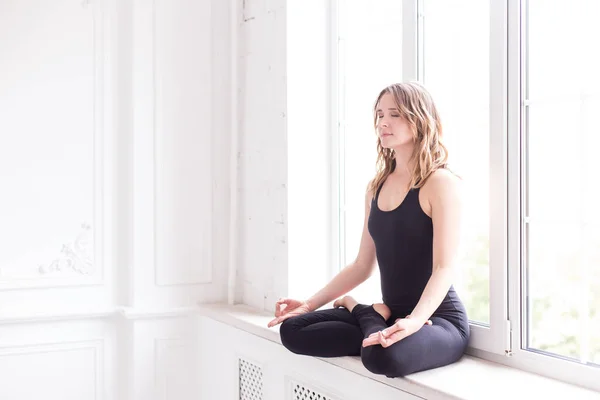 Blond kvinna i idrott outfit sitter på fönsterbrädan i Lotus pose — Stockfoto