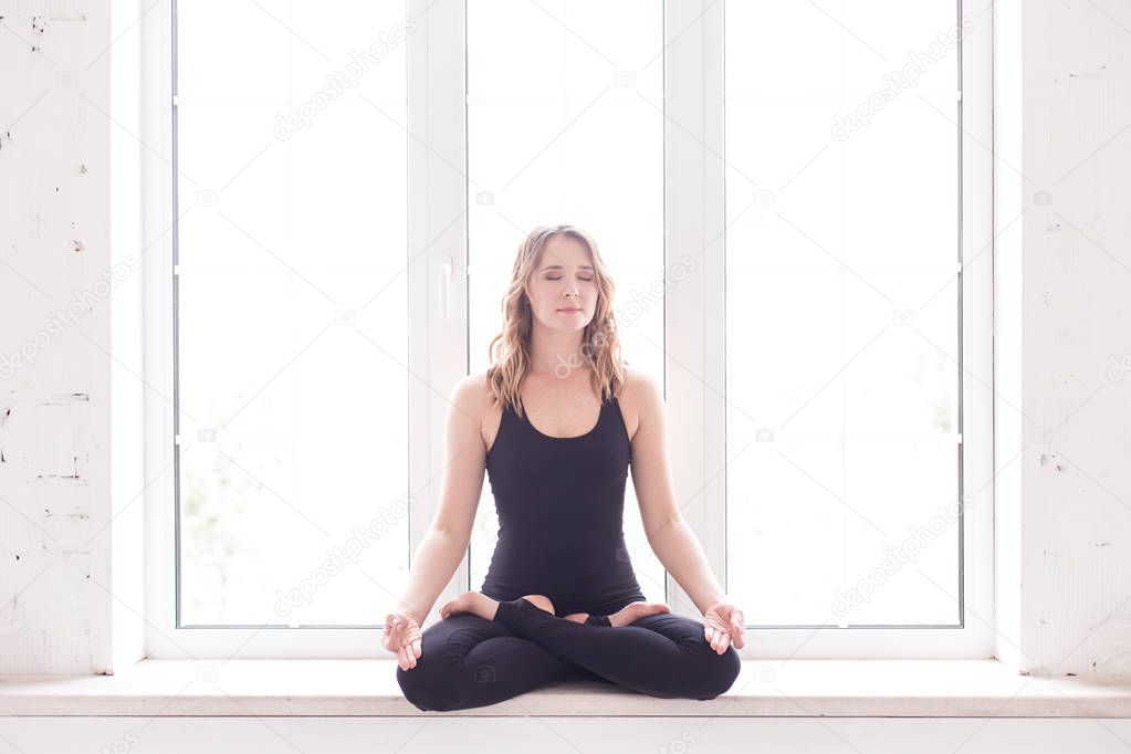 Blond woman in sport outfit sits on windowsill in lotus pose