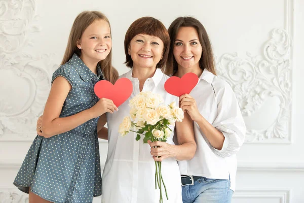 Mother, daughter and granddaughter hug and hold cardboard hearts and a bouquet of flowers