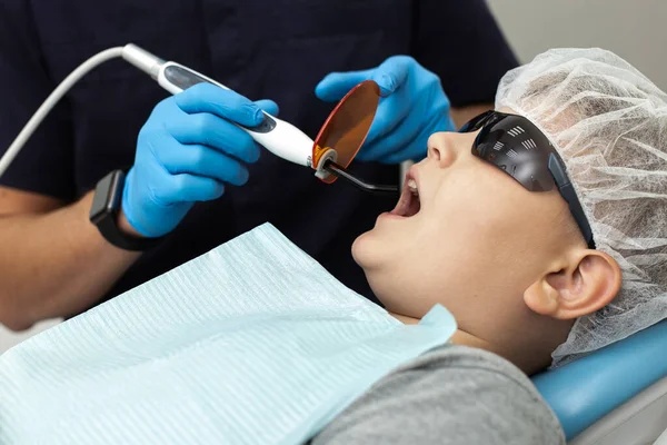 Giovane paziente in occhiali neri posa sulla sedia mentre il dentista esamina la bocca con lampada ultravioletta — Foto Stock