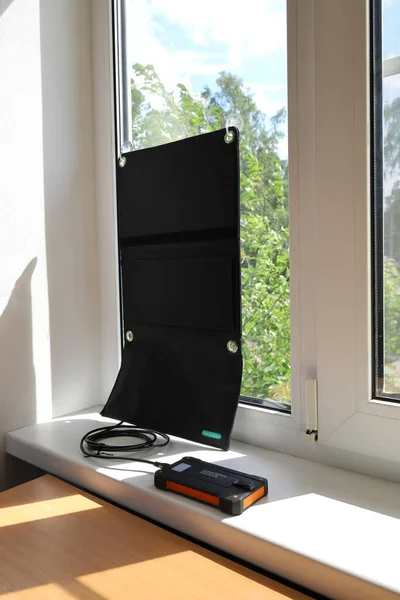 Portable solar charger on the suction cups attached to the glass of the window generates energy and charges the powerbank on the windowsill. Sunny day, vertical format