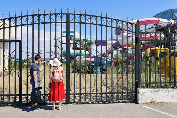 Two people stand at the closed gates of an abandoned water park and look through the bars. There are no people inside and nothing is working due to the quarantine, although the weather is good