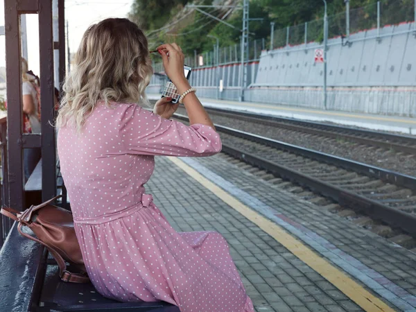 Young woman sits on bench of railway platform, waits for her train and looks in pocket mirror, corrects makeup on her face