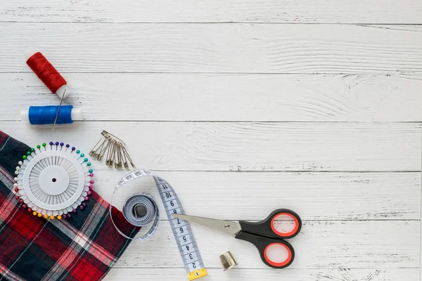 Accessoires de couture. Tissu à carreaux sur fond de bois blanc. Tissu, fils à coudre, aiguille, broches, ciseaux et centimètre de couture. vue du dessus, flatlay — Photo