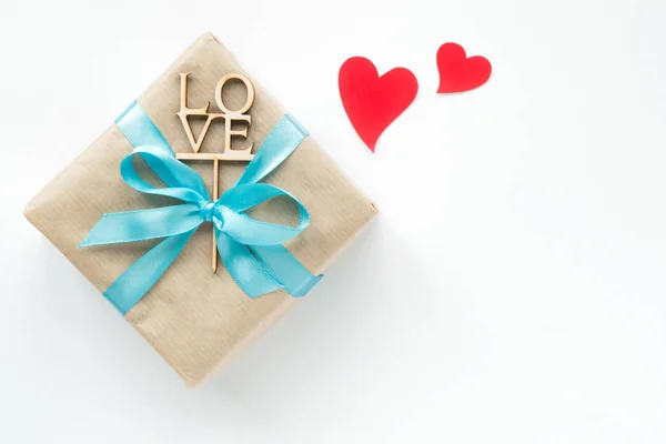 Geschenk doos gewikkeld in bruin papier met blauw lint en rode harten op witte achtergrond. Bovenaanzicht. Copyspace — Stockfoto