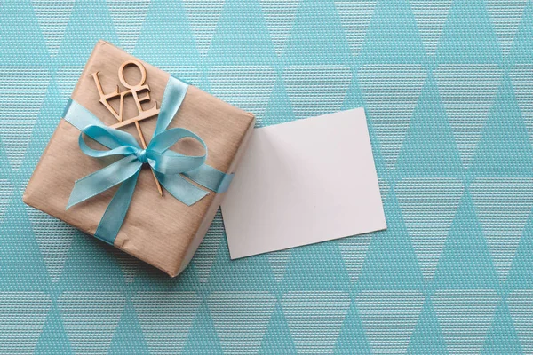 Geschenk doos verpakt in bruin papier met licht blauw lint op blauwe achtergrond met wenskaart. Mockup. Freespace — Stockfoto