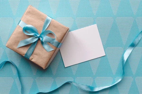 Geschenk doos gewikkeld in bruin papier met lint op blauwe achtergrond met wenskaart. Mockup — Stockfoto