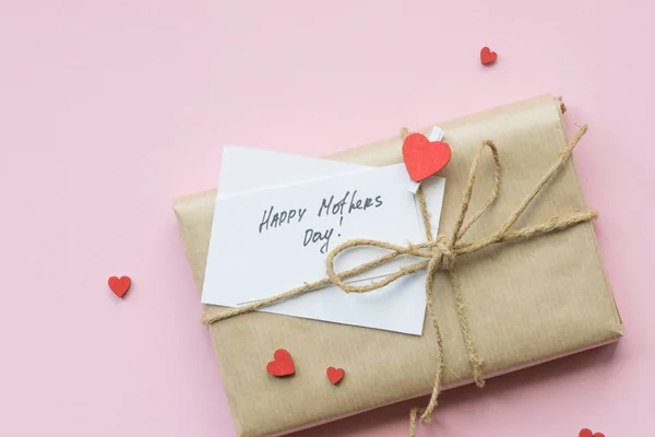 Presente avvolto in carta artigianale marrone e stringa di canapa cravatta su sfondo rosa chiaro. Confezione regalo con auguri per la Festa della Mamma. Vista dall'alto . — Foto Stock