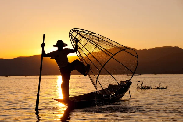 Silueta de Myanmar pescador en barco de madera al atardecer. Pescador birmano en barco de bambú captura de pescado de manera tradicional con el equilibrio de la red hecha a mano y remar con una pierna. Lago Inle, Myanmar — Foto de Stock