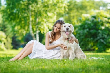 Yaz parkında Golden Retriever köpeğiyle çimenlerde oturan genç bir kadın.