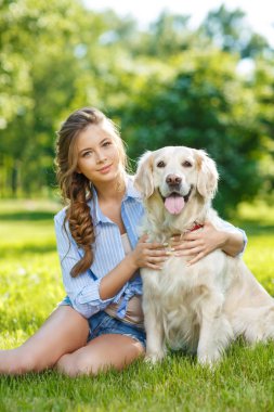 Yaz parkında Golden Retriever köpeğiyle çimenlerde oturan genç bir kadın.