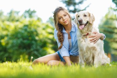 Yaz parkında Golden Retriever köpeğiyle çimenlerde oturan genç bir kadın.
