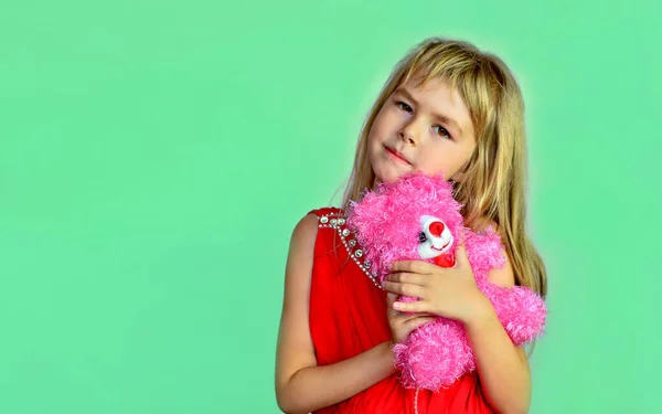 ピンクのクマの赤いドレスの少女 ターコイズの背景にピンクの柔らかいおもちゃを持つ小さな女の子 テキストのためのコピースペースを持つ茶色の目のブロンド ストックフォト