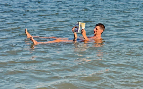 死海に浮かぶ本を読む男 死海で雑誌を読む 手に本を持って塩湖で泳いでいる 死海の水の上に男が横たわっている 塩辛い海で泳ぐ ロイヤリティフリーのストック画像