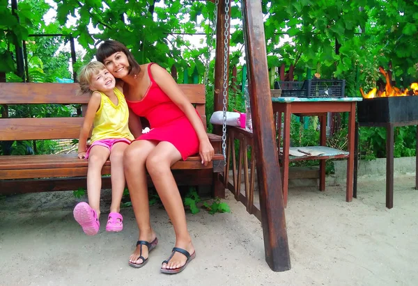 Mom and daughter cuddle cheerfully. Little girl with mom laugh fun in the garden. Family gatherings on the background of a bonfire in nature. — Stock Photo, Image