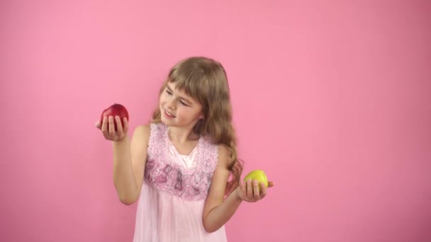 Klein meisje op een roze achtergrond met een rode appel en een groene appel. Het kind maakt een keuze tussen rode en groene appels. Een klein meisje eet fruit. Vruchtvitaminen. Moeilijke keuze. Gezond eten — Stockvideo