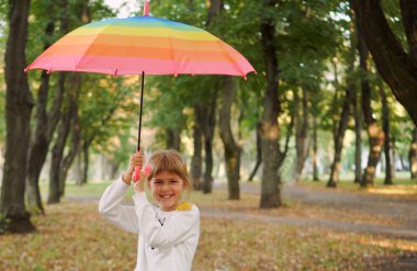 Küçük bir kız çok renkli bir şemsiyeyle neşeyle ayakta duruyor. Sonbahar yürüyüşü. Merhaba sonbahar..