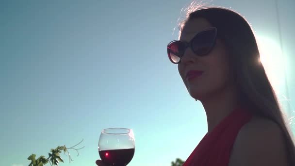 Dívka v červených šatech na pozadí modré oblohy. Dívka s dlouhými vlasy pije červené víno proti modrému nebi.