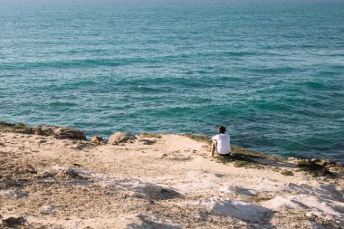 Adam uçurumun kenarında sahilde oturuyor.