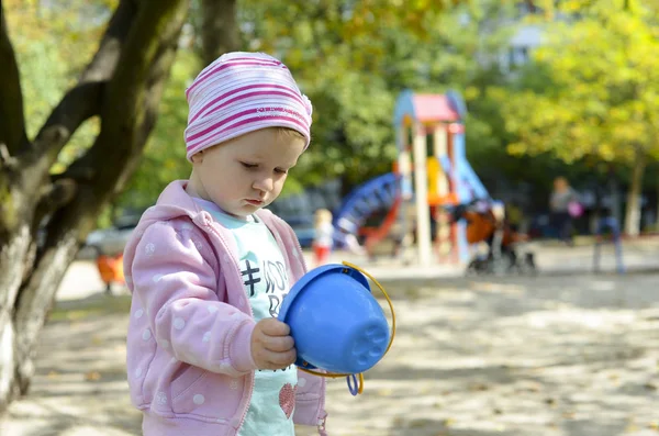 Toddler baby girl is playing outdoor and looking at the blue toy bucket. Walking with Child on the playground at sunny day, wonderful weather. Serious baby. Playing alone at kindergarten. 2, 3 years