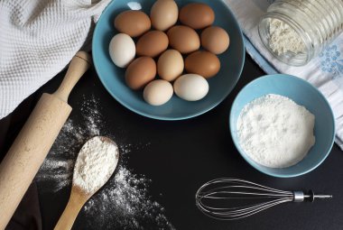 Hamurun hazırlanması. Un, yumurta, çırpma, haddeleme ve siyah masaüzerinde bir mutfak havlusu. Tahta bir kaşıkta bir demet un, mavi tabakta bir kase un ve yumurta. Pişirme maddesi üst görünümü